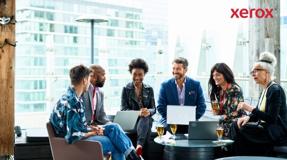 A group of seven people sit together, conversing and working on laptops with a city backdrop