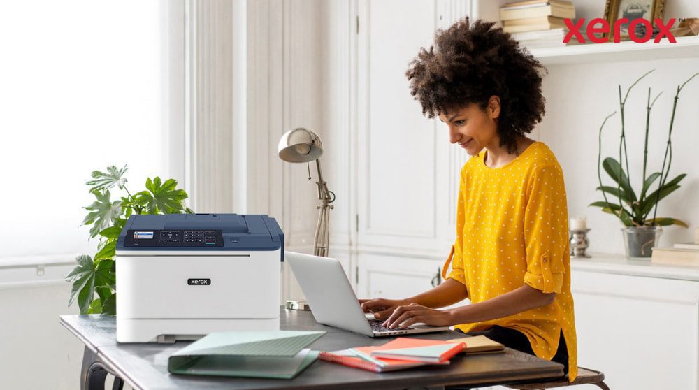 Xerox® DocuShare® Go: Productivity for Hybrid Work A person with curly hair is smiling while typing on a laptop at a desk, with a compact Xerox C310 printer positioned next to the laptop, showcasing a modern home office setup for productivity.