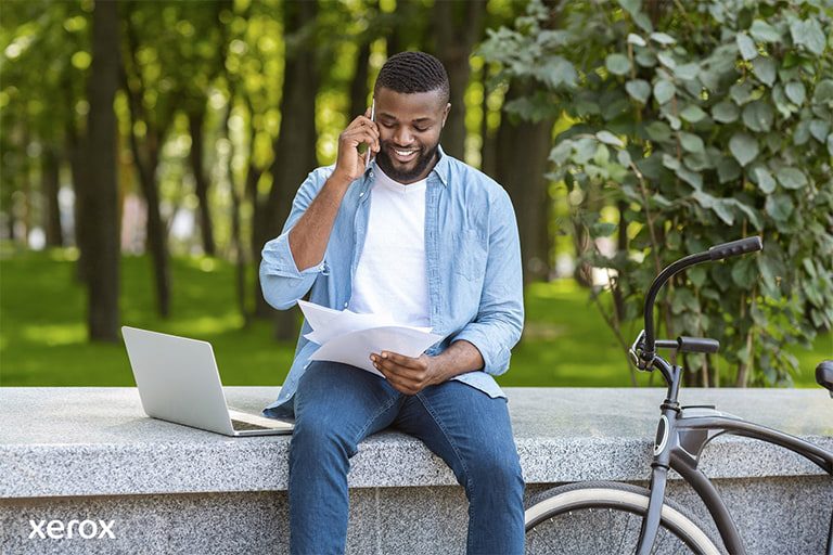 A man is seated on a bench, using his laptop, while his bicycle is parked next to him in a peaceful environment.