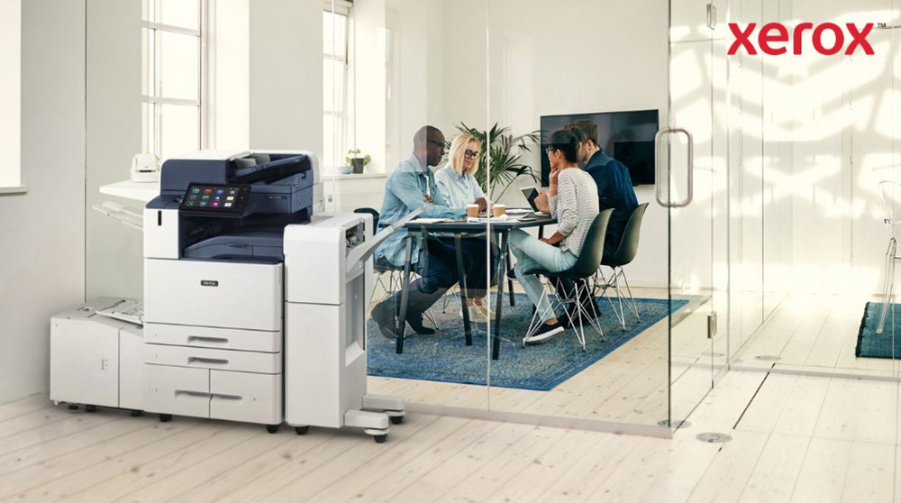 A modern Xerox AltaLink B8245 multifunction printer stands in the foreground of a bright office. In the background, a group of people are engaged in a collaborative discussion around a table.