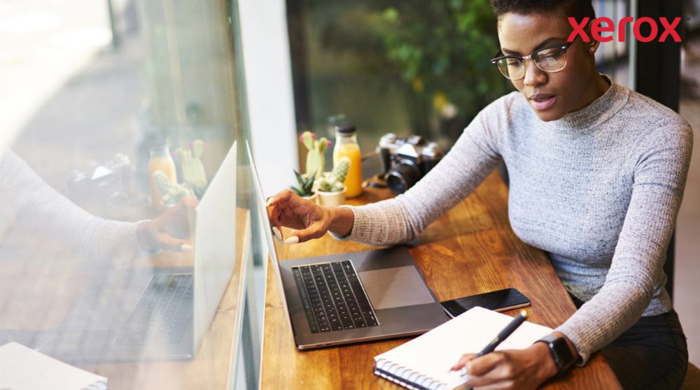 Xerox® Workplace Solutions: Enhanced Productivity A person wearing glasses is working at a wooden table, looking at a laptop screen and writing in a notebook, with a smartphone nearby, suggesting a productive work environment